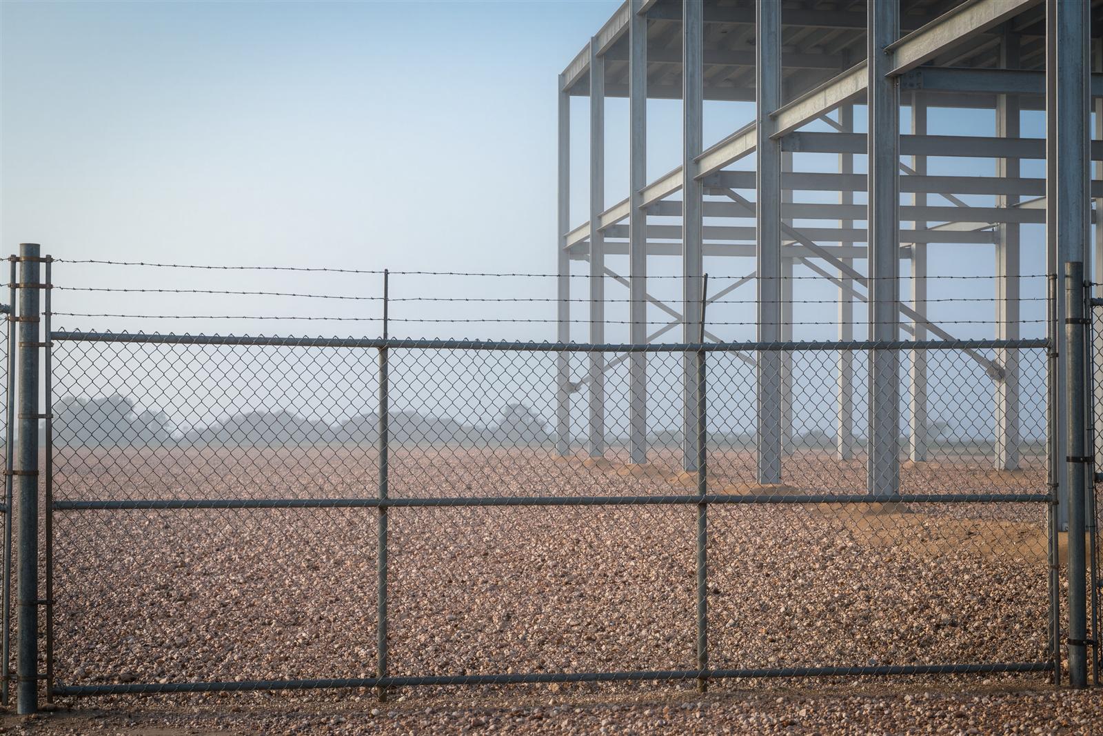 A chain link construction gate with barbed wire in front of a steel frame building rising in morning fog on a gravel lot