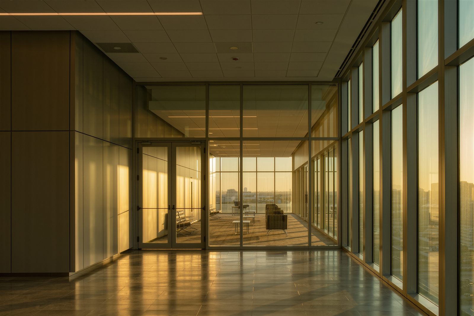 A quiet office corridor with a glass entry door and lobby seating lit by warm evening light through tall windows