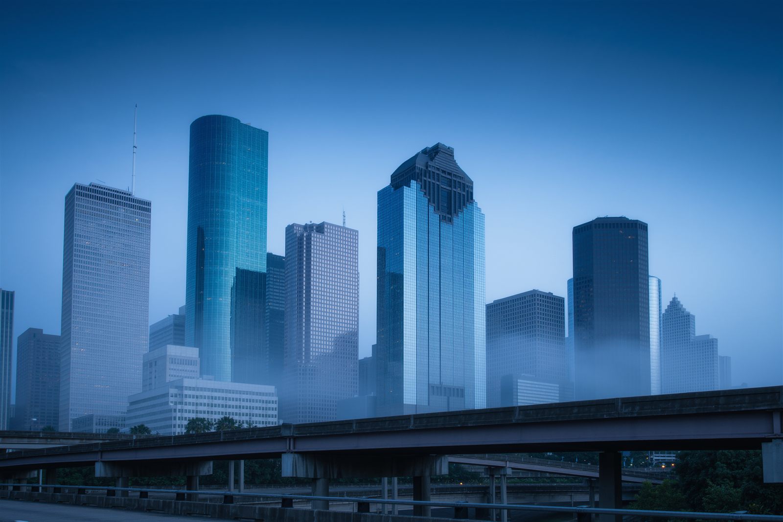 A cluster of glass downtown office towers at blue hour seen from behind a freeway overpass in light haze