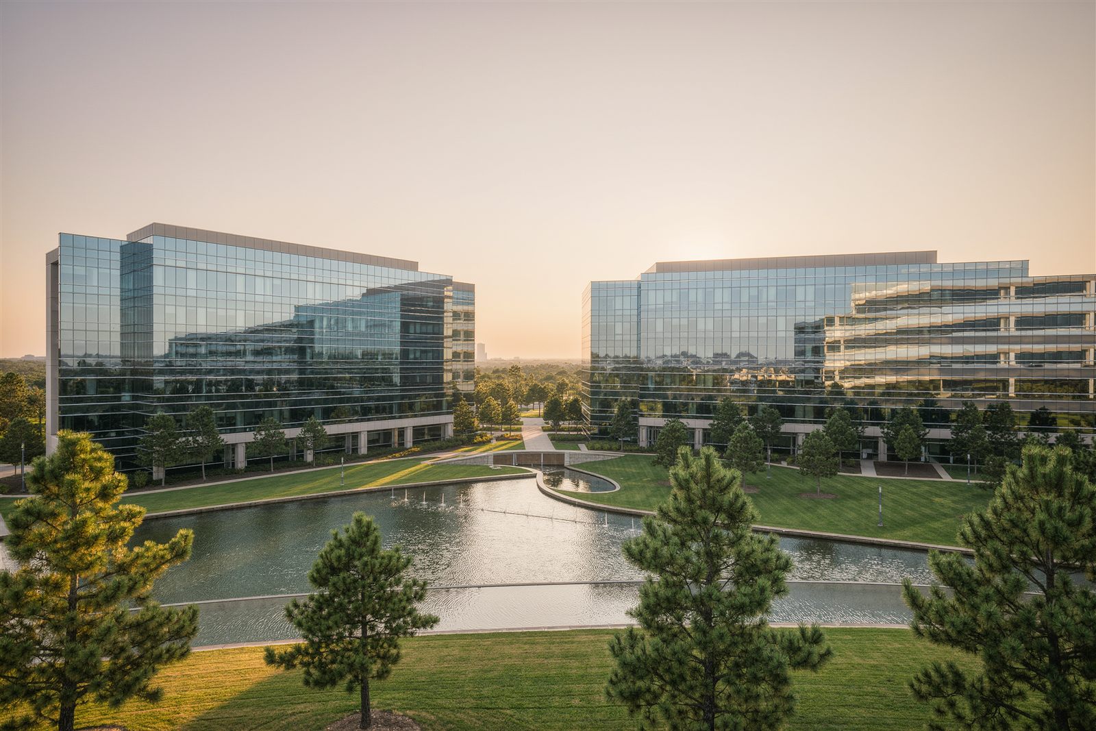 Low glass office buildings beside a landscaped pond and pine trees in a suburban office park at sunrise