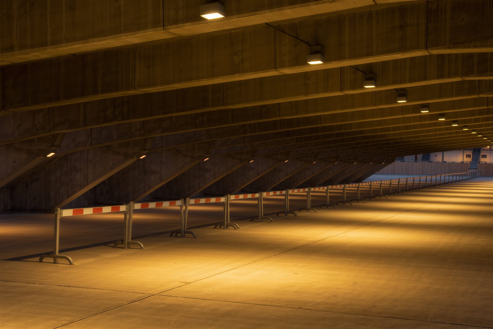 A long empty stadium concourse at night with red and white crowd barriers lined up under concrete ceiling lights