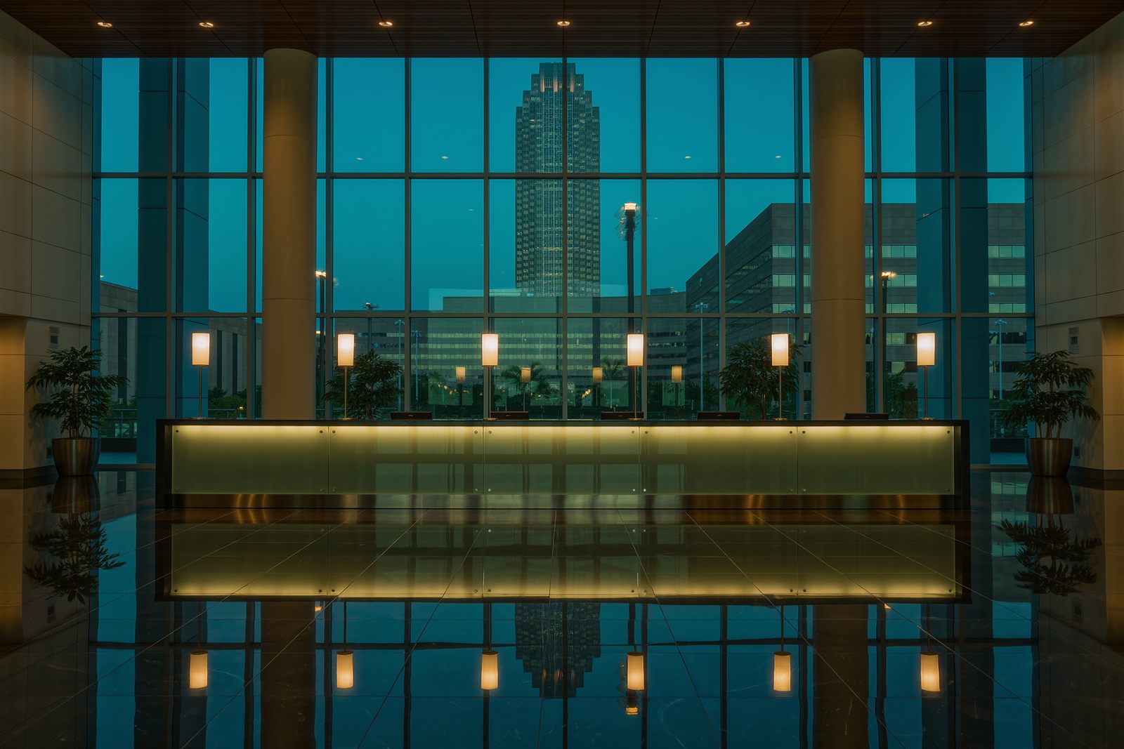 An empty glass office lobby at dusk with a long reception desk, warm lamps and tall windows looking out over a city tower