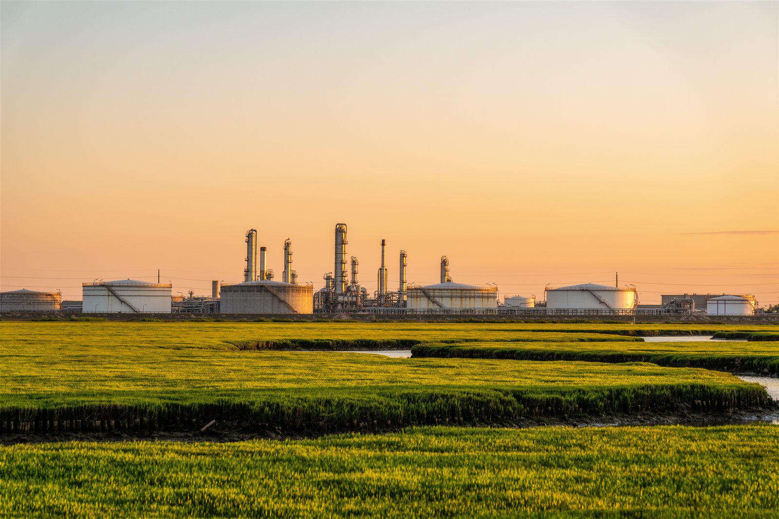 A line of white storage tanks and refinery towers across green marsh grass and a quiet channel at sunset