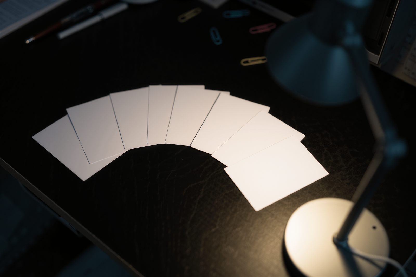 Several blank white cards fanned out on a dark desk under a desk lamp with paper clips scattered nearby